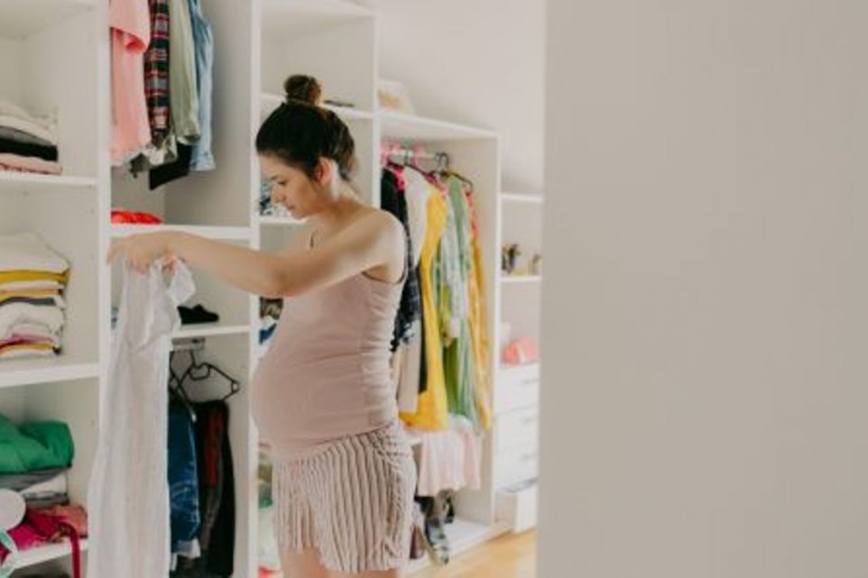 Photo of an expectant mother folding and organizing clothes in the dressing room of her apartment; the daily routine of a young pregnant woman.