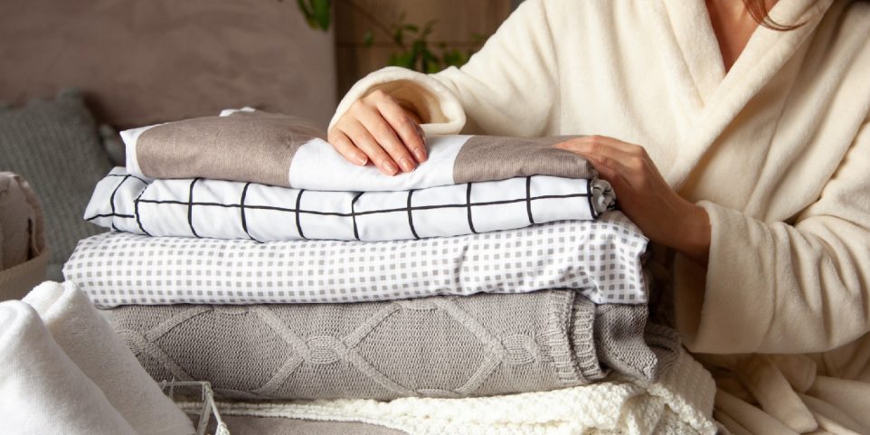 Beautiful woman in winter thick warm robe is sitting and neatly folding bed linens and white bath towels.