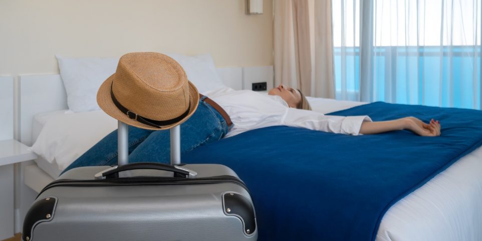 Close-up of a gray suitcase with a hat on the background of a young woman in blue jeans and a white shirt lying on the bed with her arms outstretched in the room. Concept of travel, recreation