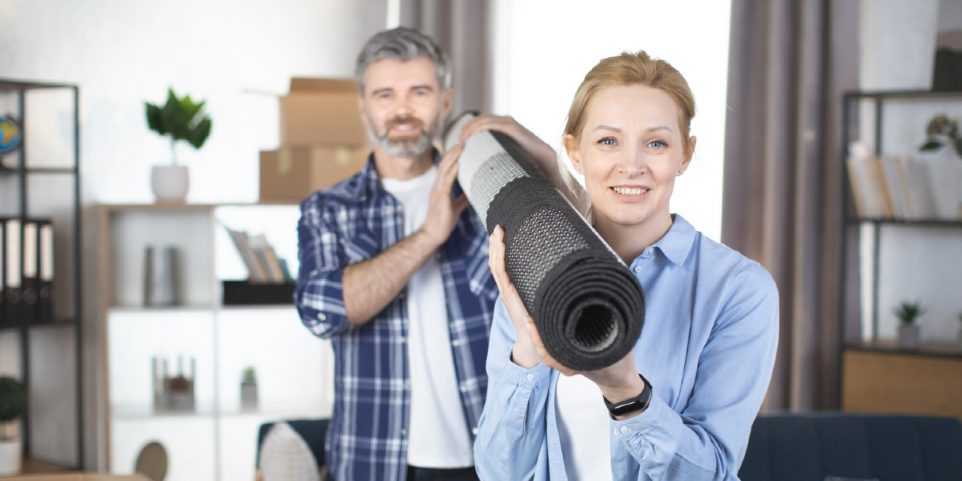 Focus on smiling pretty blond woman, holding roll carpet on her shoulder and her pleasant bearded man behind